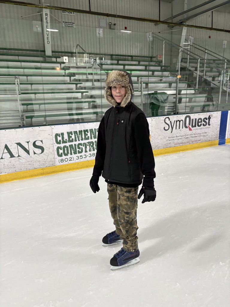 A student wearing a winter hat and gloves stands on an indoor ice rink in skates, posing near the boards with empty bleachers in the background.