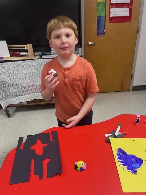 A young boy with strawberry-blonde hair wearing an orange henley shirt stands in a classroom. He holds a small, colorful craft made of red and white pom-poms with googly eyes. On the red table in front of him are scraps of black construction paper and another yellow pom-pom creature.