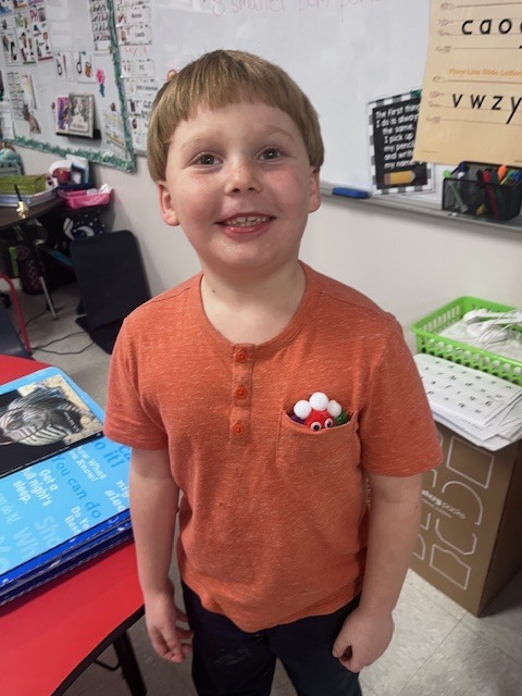 A close-up portrait of the boy in the orange henley shirt, smiling happily. He has tucked his red and white pom-pom creature into his shirt’s chest pocket so only its eyes and the top of its "head" are visible.