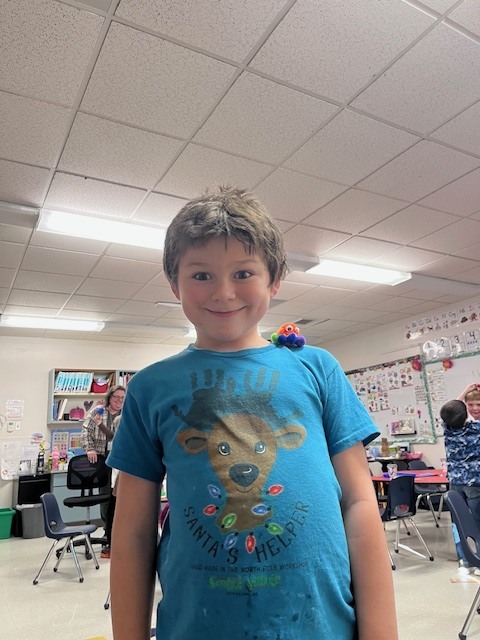 A young boy in a bright blue t-shirt featuring a reindeer "Santa's Helper" graphic smiles broadly for the camera. A small, colorful pom-pom creature with googly eyes is perched playfully on his left shoulder.