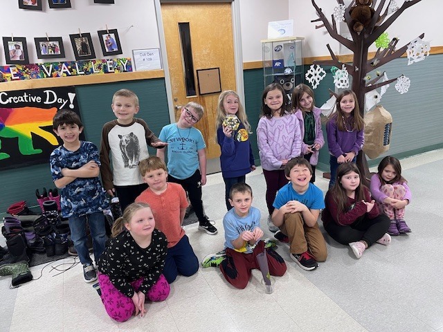 A group portrait of twelve elementary-aged students posing together in a school hallway. They are kneeling and standing in front of a wall decorated with student photos and a paper "Creative Day" display. Many of them are holding or wearing their pom-pom crafts.