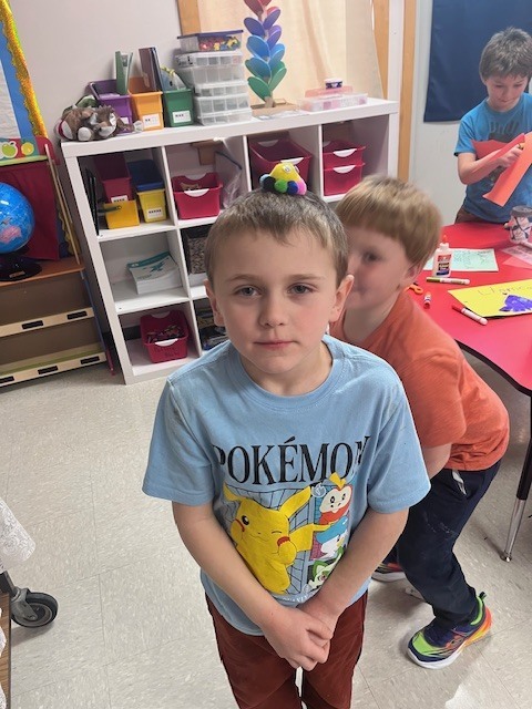 A young boy in a light blue Pokémon t-shirt stands in a classroom with a neutral expression, balancing a large, multicolored yarn pom-pom with googly eyes on top of his head. In the background, another child in an orange shirt sits at a red table, smiling. The classroom is filled with educational toys, including a globe, a colorful stacking tower, and storage bins.