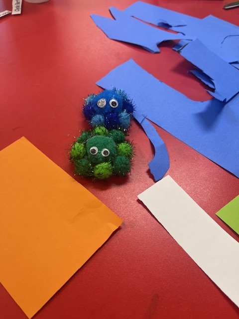 A close-up of two pom-pom creatures sitting on a red table. One is blue and sparkly, and the other is green. They are surrounded by scraps of blue, orange, and white construction paper, showing a work-in-progress art project.