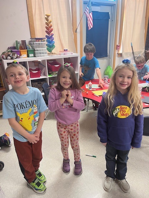 Three children—a boy in a blue Pokémon shirt, a girl in a pink sweatshirt, and a girl in a purple Bass Pro Shops hoodie—stand in a line, smiling. Each child has a small pom-pom creature balanced on their head like a tiny hat.