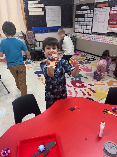 A young boy in a blue camouflage-patterned shirt smiles as he holds up a craft made of tan paper and several multicolored pom-poms. He is standing in a classroom with a colorful alphabet rug in the background.