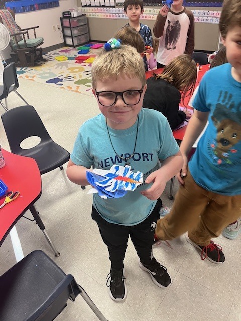 A boy with glasses and a light blue "Newport" t-shirt holds a colorful paper craft. He has a blue and green pom-pom creature balanced on top of his head. Other children are visible in the background of the classroom.
