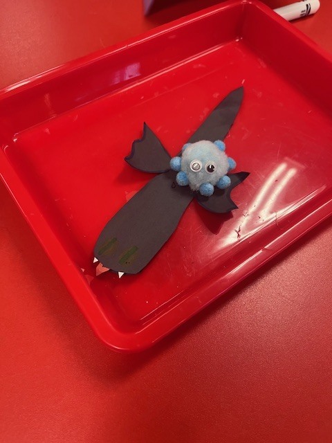 A top-down close-up of a craft project resting in a red plastic tray. The craft consists of a blue pom-pom creature with googly eyes attached to a black construction paper cutout shaped like a bat or a stylized bird.