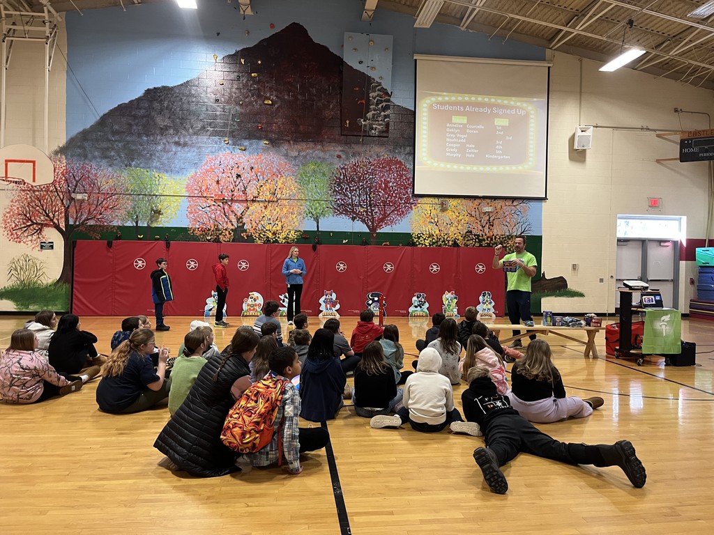 Elementary students sit on the gym floor at Castleton Elementary School during the Kids Heart Challenge kickoff assembly while a presenter speaks at the front, with colorful heart-themed characters displayed and a mural painted on the gym wall behind them.