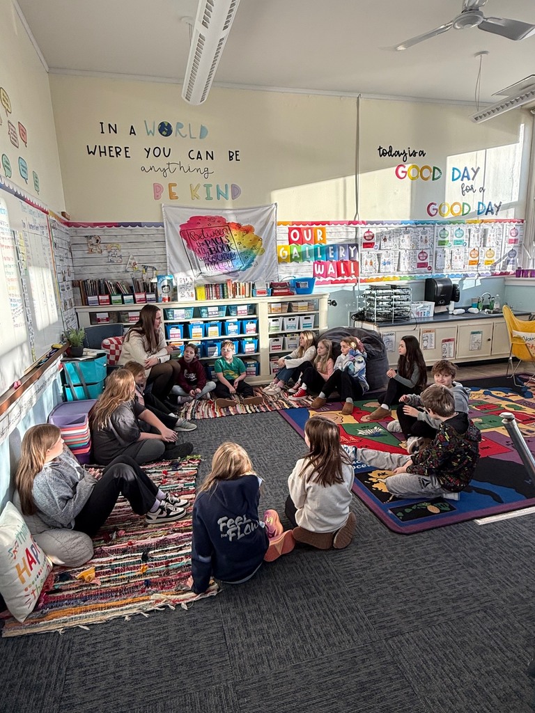 An elementary school classroom scene shows a group of about twelve children and one adult teacher sitting in a large circle on the floor. They are gathered on colorful rugs, including a large map rug of the United States. The room is brightly lit by natural light and features several inspirational wall displays