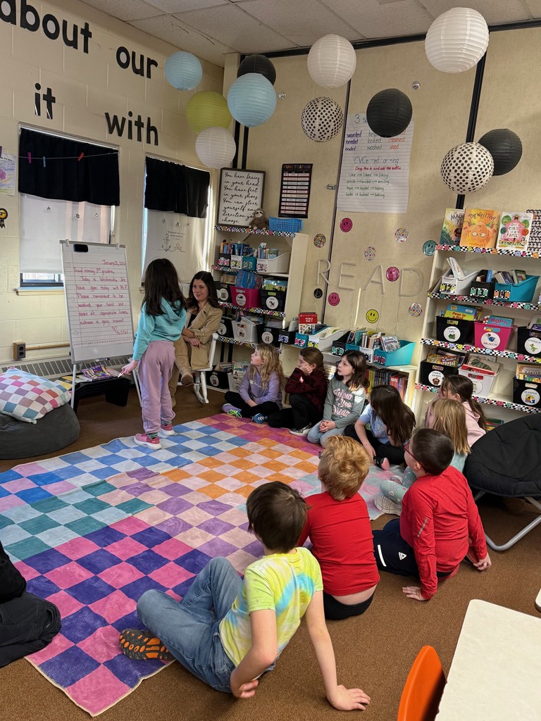 Elementary students sit on a colorful rug in a reading corner while a teacher listens to a child standing and speaking during morning meeting, with bookshelves and hanging lanterns in the background.