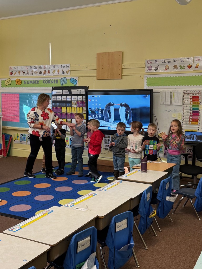 A classroom setting showing a teacher and seven young children standing in a row in front of a large digital display. The screen shows an image of two penguins, and several students appear to be using hand gestures. The room includes a "NUMBER CORNER" display, an alphabet line, and student desks with blue chair pockets
