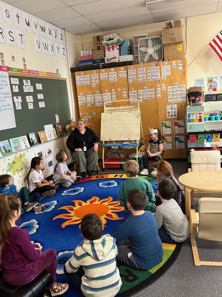Teacher and young students sit in a circle on a classroom rug during a group discussion, surrounded by alphabet charts, books, and learning materials.