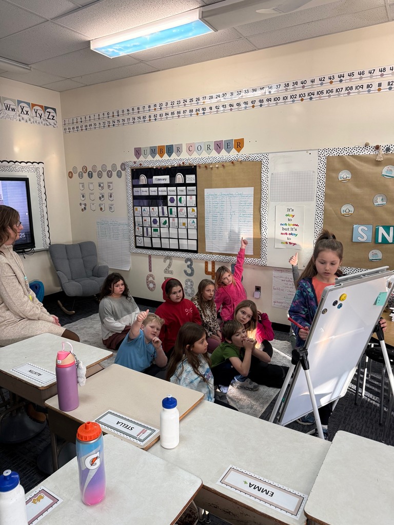 An elementary school classroom scene where a young girl stands at a white easel, leading a lesson for a group of seven children seated on a rug. An adult teacher sits nearby in a chair, observing. The background features educational posters including a "Number Corner," a calendar, and a number line. I