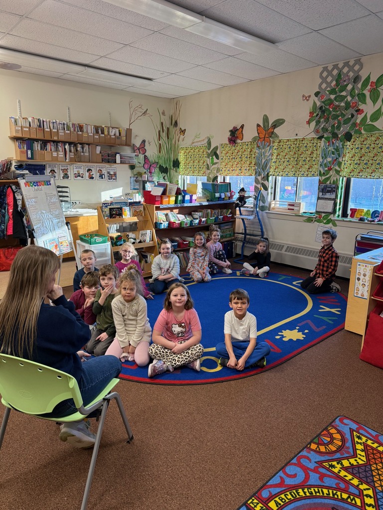 Students sit in a circle on a classroom rug facing their teacher during morning meeting, with bookshelves and butterfly wall murals in the background.