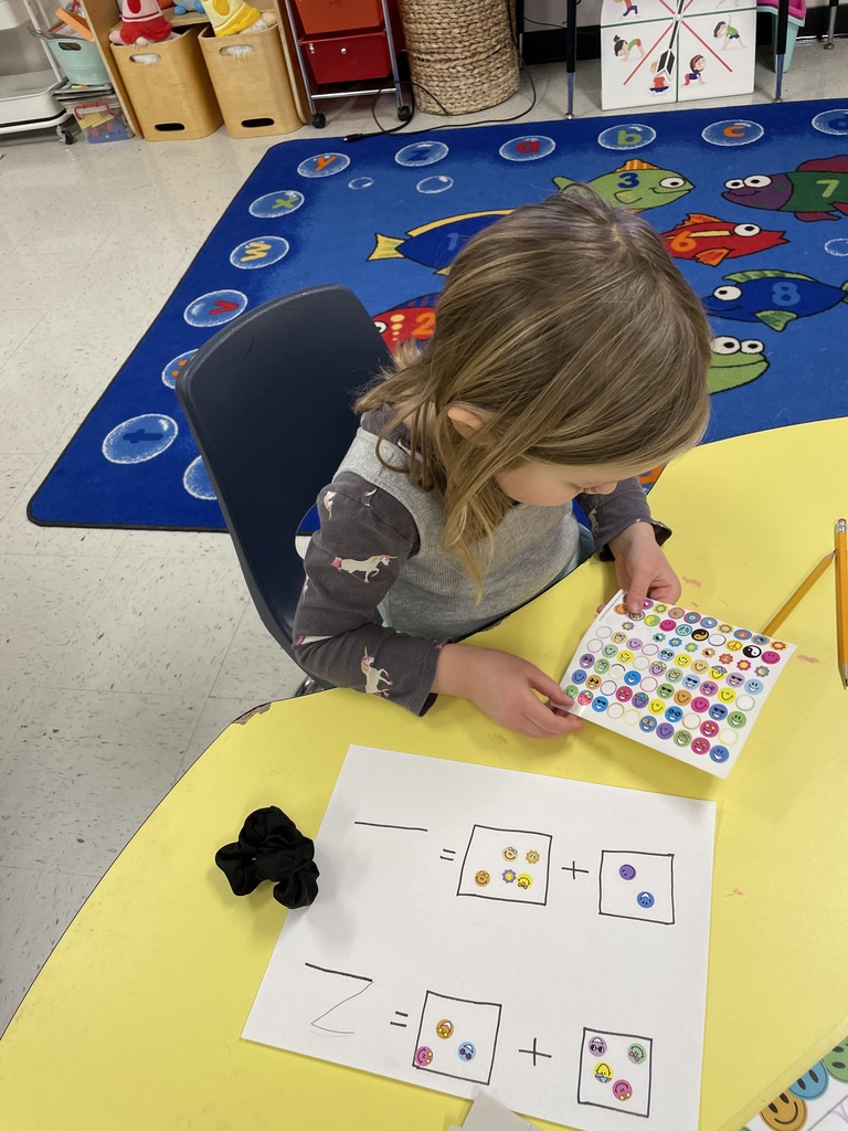 A high-angle shot of a young girl with blonde hair in a grey and purple unicorn-patterned shirt. She is focused on peeling a sticker from a sheet to place onto her math worksheet. A black hair scrunchie sits on the table beside her paper. IMG_5297.png: An overhead view of a child with blonde hair working at a blue table. The child is holding a sticker sheet over a worksheet that shows two addition problems: one totaling seven and one totaling five, represented by sticker groups.