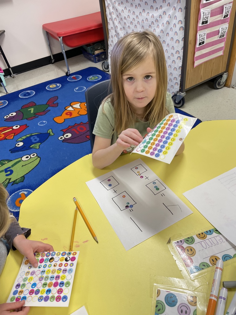 A young girl with long blonde hair looks toward the camera while holding a sheet of colorful smiley-face stickers. In front of her is a math worksheet with hand-drawn squares and addition symbols.