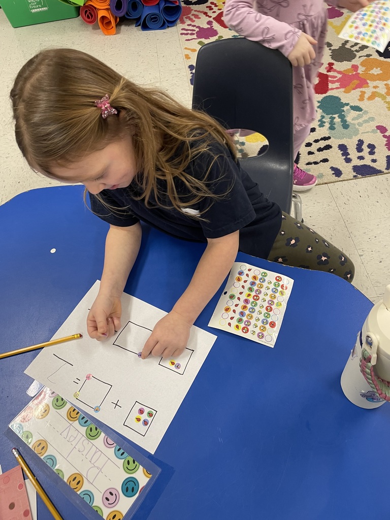 A young girl with a pink butterfly clip in her hair leans over a blue table. She is using her fingers to precisely place a small ladybug sticker into a box on her math assignment.