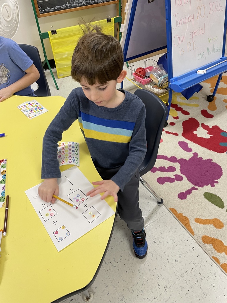 A young boy with short brown hair wearing a blue, yellow, and white striped long-sleeve shirt sits at a yellow table. He is using a yellow pencil to point at a math worksheet featuring boxes filled with colorful stickers and addition signs.