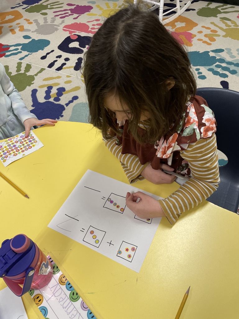 A young girl with dark wavy hair wearing a floral and striped rust-colored dress looks down at her math paper. She is carefully placing stickers into the designated boxes to complete her addition lesson. Overhead and Action Shots