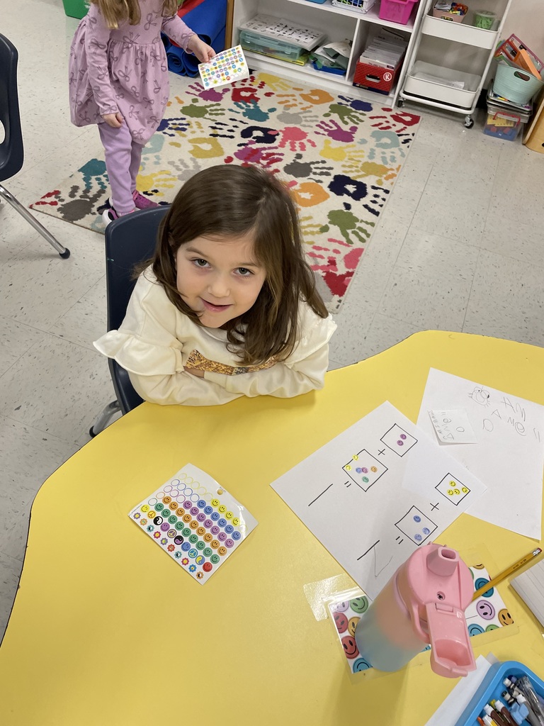 A young girl with brown hair and a cream-colored ruffled shirt smiles at the camera. She sits at a yellow table with a math worksheet and a sheet of stickers in front of her.