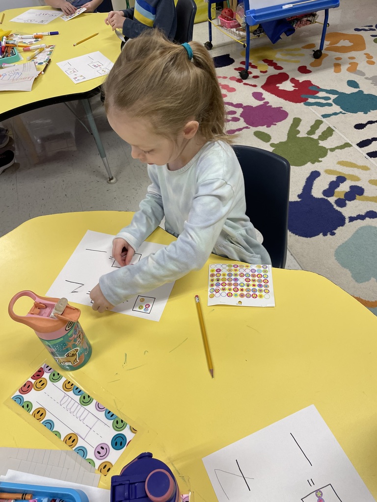 A young girl with blonde hair in a ponytail and a light blue long-sleeve shirt works on her math. She is mid-action, placing stickers into the boxes on her paper. A colorful name tag is visible on the table. Classroom Environment Background Details: Several photos show a vibrant classroom s