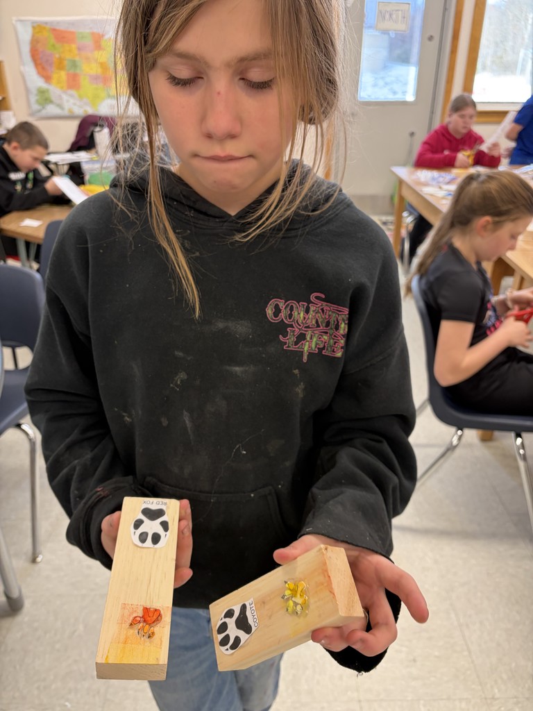 A student stands in a classroom holding up two finished wooden stamps. One stamp features a "Red Fox" track label and the other a "Coyote" track label, both with 3D elements attached for stamping.