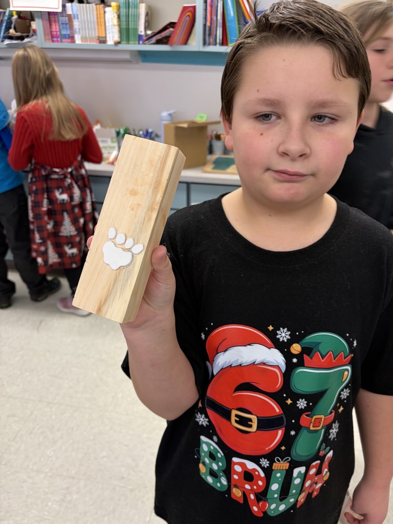A student in a black holiday-themed t-shirt holds up a long wooden block with a white foam animal paw print glued to the surface, showing the progress of their handmade stamp.