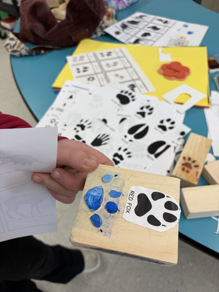A close-up of a student holding a square wooden block. They have glued blue foam pieces onto the wood to mirror the "Red Fox" track printed on a paper guide next to it.