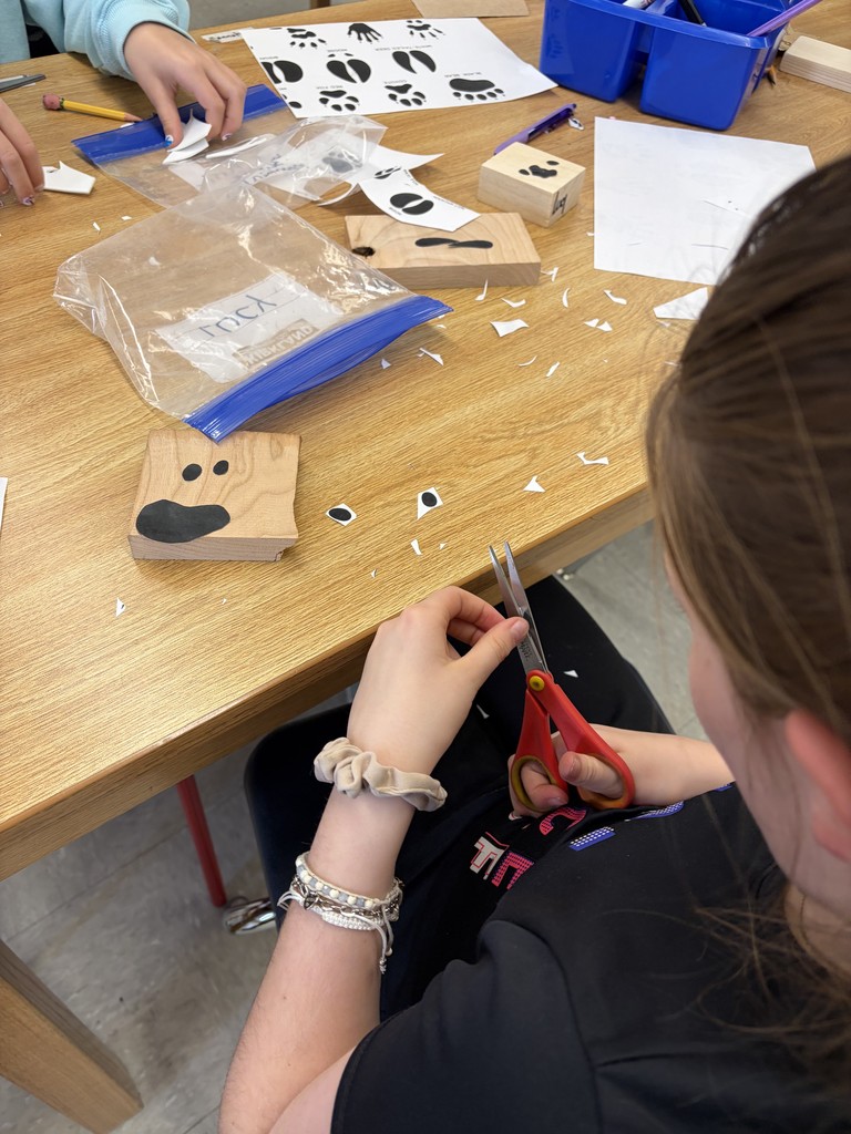 A close-up view of two students at a workstation using hot glue guns to attach blue foam animal track shapes onto rectangular wooden blocks.