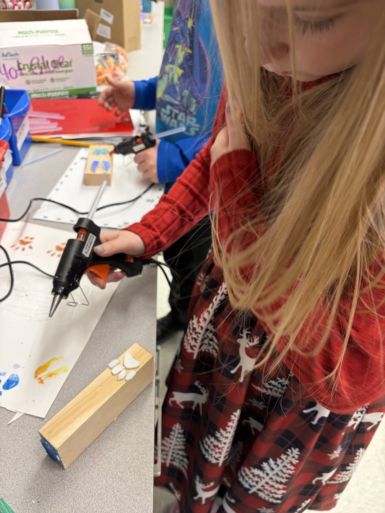 A student in a red plaid dress uses a hot glue gun to secure a white foam paw print onto a wooden block. In the background, another student works on a similar project at the same craft station.