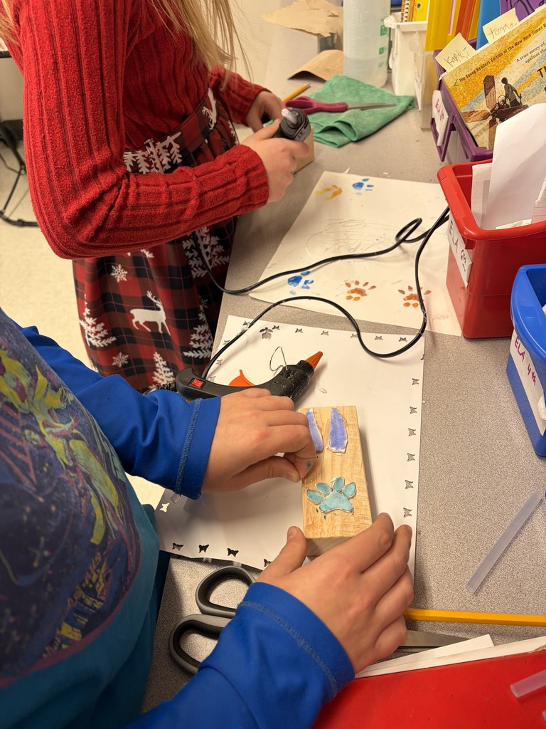 A close-up view of two students at a workstation using hot glue guns to attach blue foam animal track shapes onto rectangular wooden blocks.