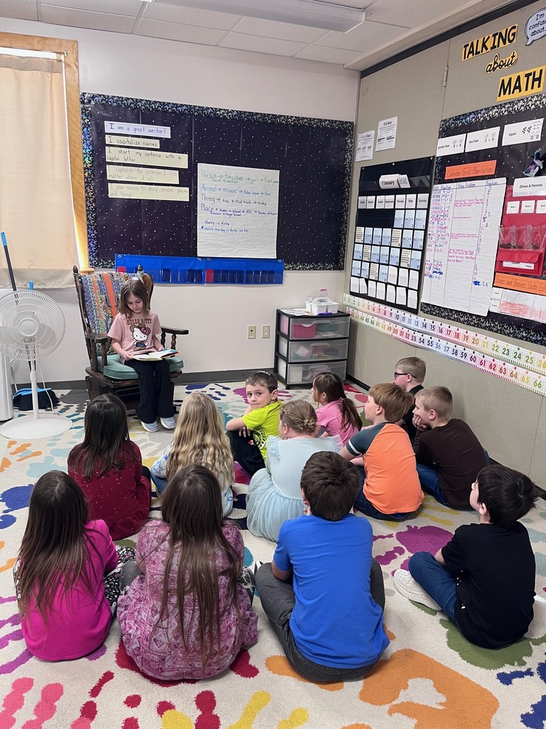 An elementary classroom scene where a group of young students sit cross-legged on a colorful rug, listening as one student sits in a rocking chair and reads from a book. The walls display anchor charts about writing and math, a calendar, and number lines. The atmosphere is calm and focused during a classroom reading or sharing time.