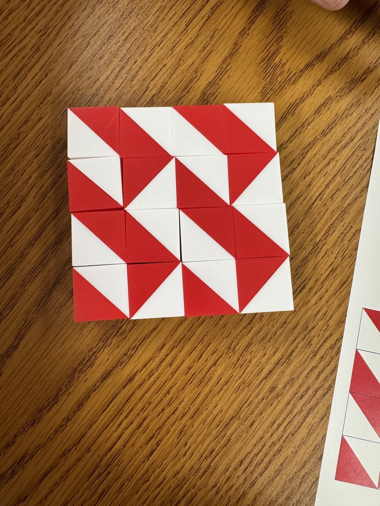 A top-down view shows a $3 \times 4$ grid of red and white blocks resting on a wooden tabletop. Each block is a square divided diagonally into a red half and a white half. The blocks are arranged to form a repeating, jagged pattern of red stripes and shapes. In the bottom right corner, a portion of a white paper is visible, showing a printed reference diagram for the pattern.