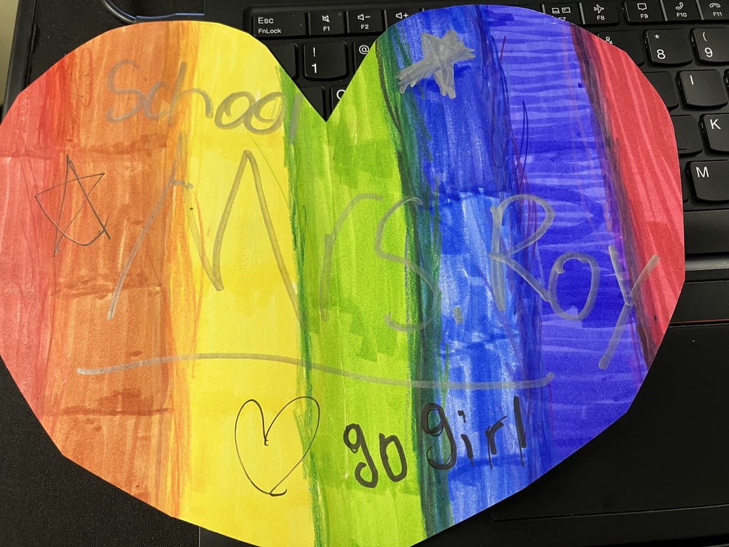 A child's drawing on a heart-shaped piece of paper featuring vertical rainbow stripes. The name "Mrs. Roy" is written in silver marker across the middle. Additional text includes "School" at the top and "go girl" at the bottom, alongside hand-drawn stars and a small heart.