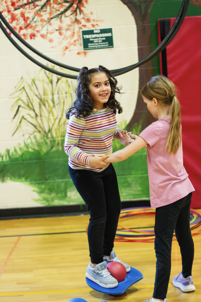 Two students smiling and helping each other balance on a wobble board during circus activities.