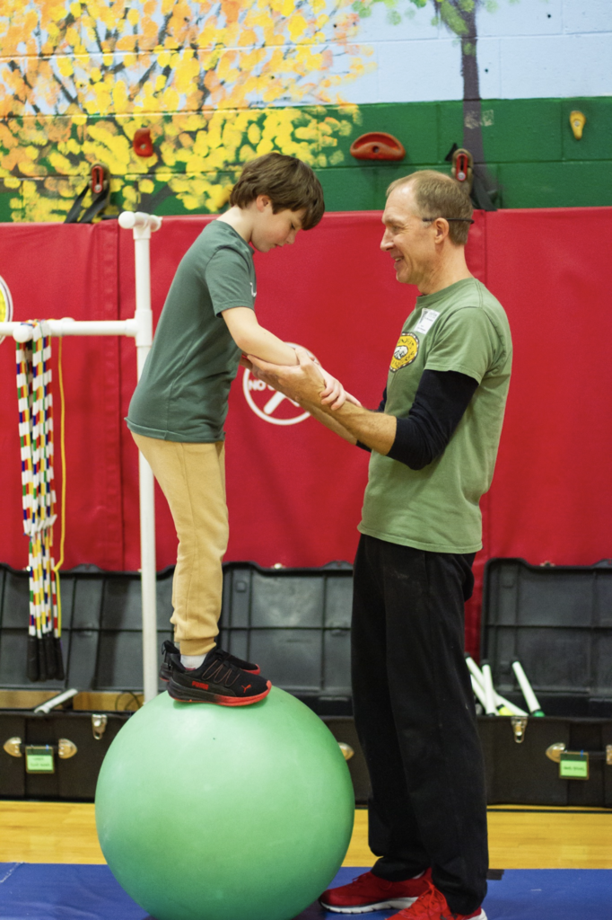 Instructor supporting a student standing on a large balance ball during a supervised circus exercise.