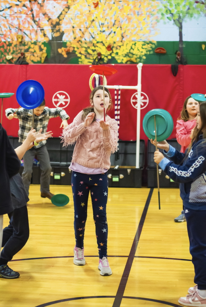 Student spinning a plate on a stick as classmates watch during a circus activity in the gym.