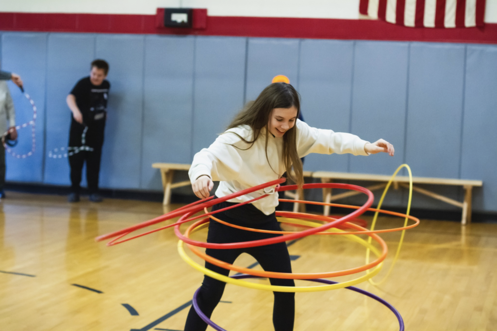 Student hula-hooping with multiple colorful hoops during a circus skills activity in the school gym.