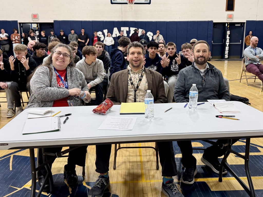 Three judges sit at a table in the gym with papers and water bottles, smiling as students and audience members gather behind them.