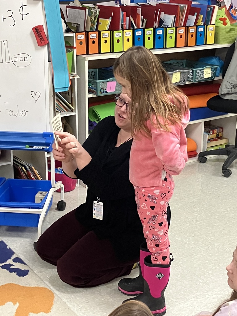 An adult kneels on the classroom floor at a child’s eye level, holding up a small card and reading or explaining it to a young child who stands beside them. The child wears pink clothing and boots. Behind them are classroom shelves with labeled bins, folders, and learning materials, creating a warm, organized early elementary classroom setting.