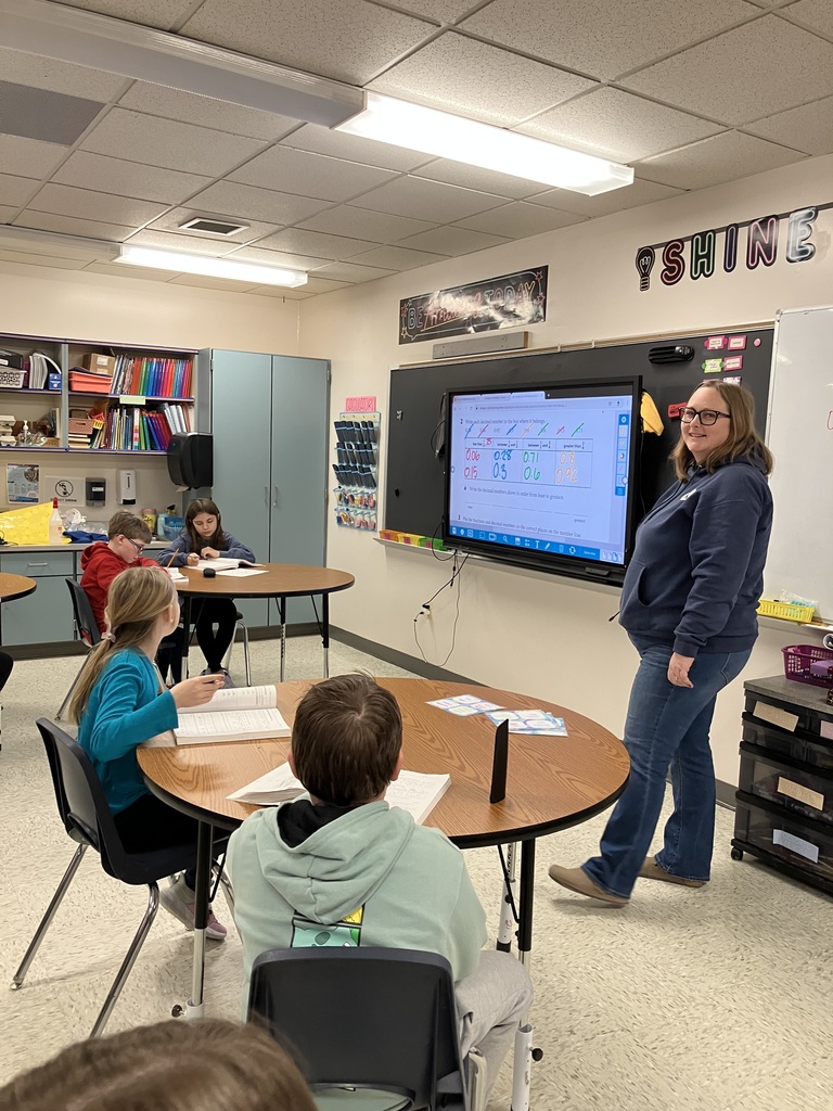 A teacher stands at the front of an elementary classroom beside a large interactive screen displaying a math lesson with decimals. Several students sit at round tables working in notebooks and looking toward the screen. The room has cabinets, bookshelves, and classroom supplies, with colorful posters on the walls and bright overhead lighting.