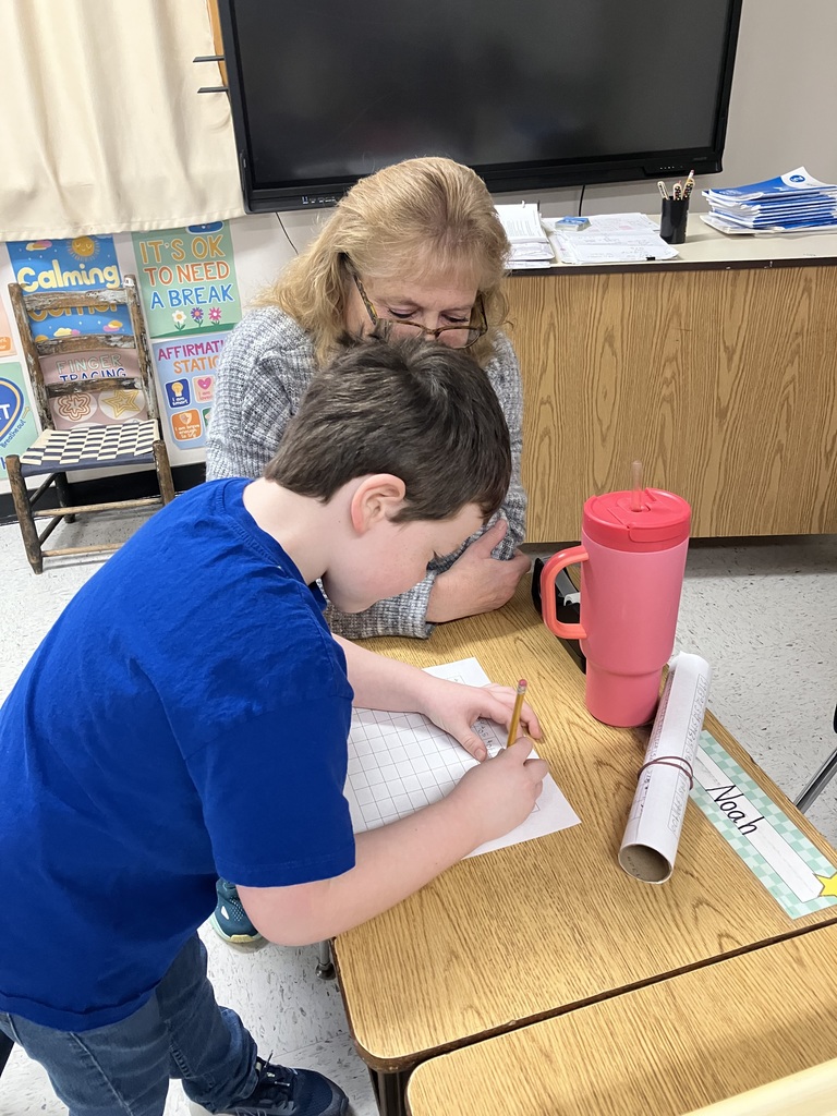An adult sits beside a student at a classroom table, closely supporting the child as they write on graph paper with a pencil. The student leans forward, focused on their work. On the table are a pink water bottle, rolled paper, and classroom materials. In the background are posters promoting calming strategies and affirmations, along with a large screen and teacher workspace, creating a supportive learning environment.