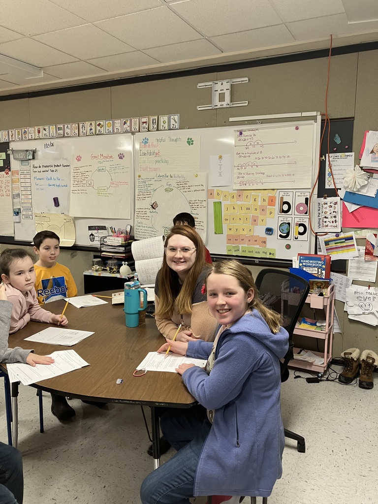 In this classroom scene, an adult woman (likely a teacher) and several young children are gathered around a kidney-shaped table. The woman, wearing glasses and a tan sweater, smiles warmly at the camera, as does a young girl in a blue fleece jacket who is holding a pencil over her worksheet. Two other students—a boy in a yellow Reebok shirt and a girl in a pink top—are also seated at the table, engaged in their work.