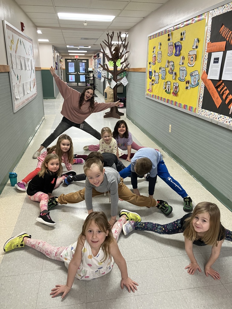 An adult woman and a group of young children are playfully posing for a photo in a school hallway, all attempting to do the splits or wide-legged stretches. The woman stands in the background with her arms and legs outstretched, while about nine children are arranged on the floor in front of her, mirroring the pose with varying degrees of success. Everyone is smiling or looking toward the camera. The hallway has green wainscoting and is decorated with colorful bulletin boards showcasing student work.