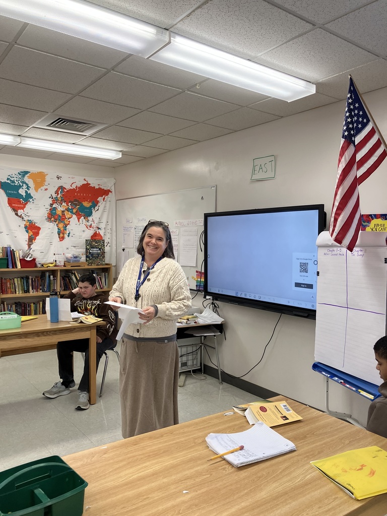 A teacher stands at the front of an elementary classroom beside a large interactive screen with materials in their hands. Several students sit at round tables writing and looking toward the teacher. The room has cabinets, bookshelves, and classroom supplies, with colorful posters on the walls and bright overhead lighting