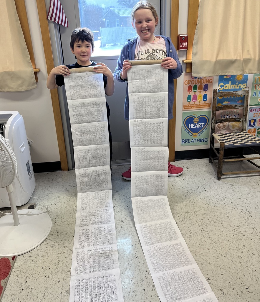 Two elementary-aged students stand indoors in front of a door, smiling and holding very long strips of paper that extend from their hands down to the floor. Each strip is covered with neatly written numbers or math work in small boxes, showing an extensive completed activity. The student on the left wears a dark shirt, and the student on the right wears a light shirt with a hoodie. Classroom posters about calming strategies and breathing are visible on the wall to the right, and a fan sits on the floor to the left.