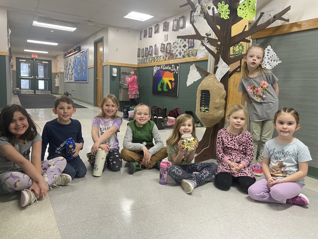 "A group of eight young children sitting on the floor in a school hallway, smiling at the camera. They are in front of a large, craft-style tree with a beehive and paper bees attached. One child is holding a jar filled with small yellow and white balls. Behind them, a bulletin board displays a colorful bear with the words 'Have a Creative Day!' and various student photos. In the background, another child in a pink outfit is at a water fountain near the hallway doors."