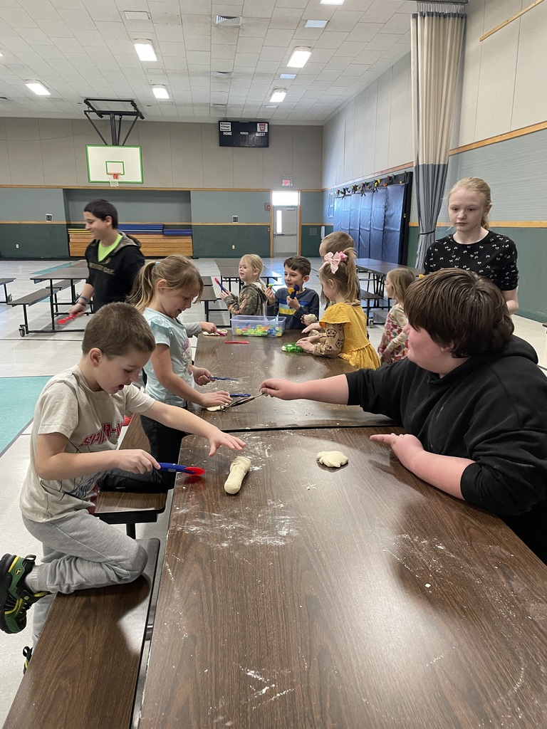 This image shows a group of children and teenagers gathered around long tables in what appears to be a school cafeteria or gymnasium, enthusiastically working with dough.