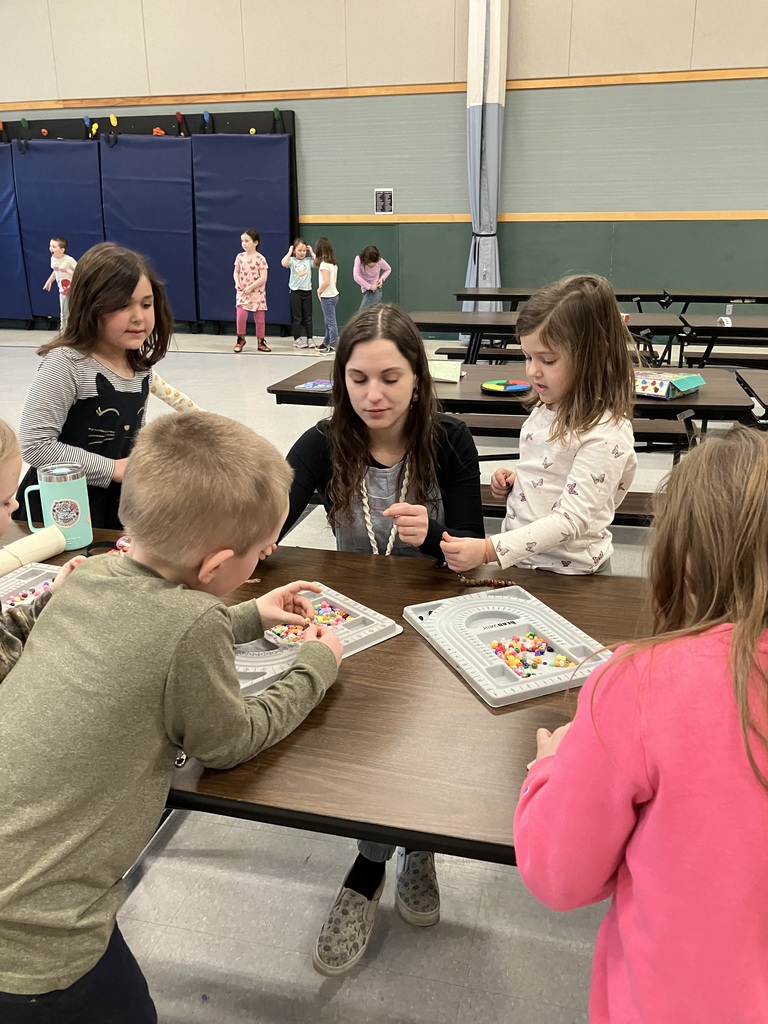 A woman and four young children sit at a long brown table in a gymnasium, focused on making jewelry with colorful beads and grey sorting trays. Several other children are visible in the background near a blue padded wall and folding cafeteria tables.