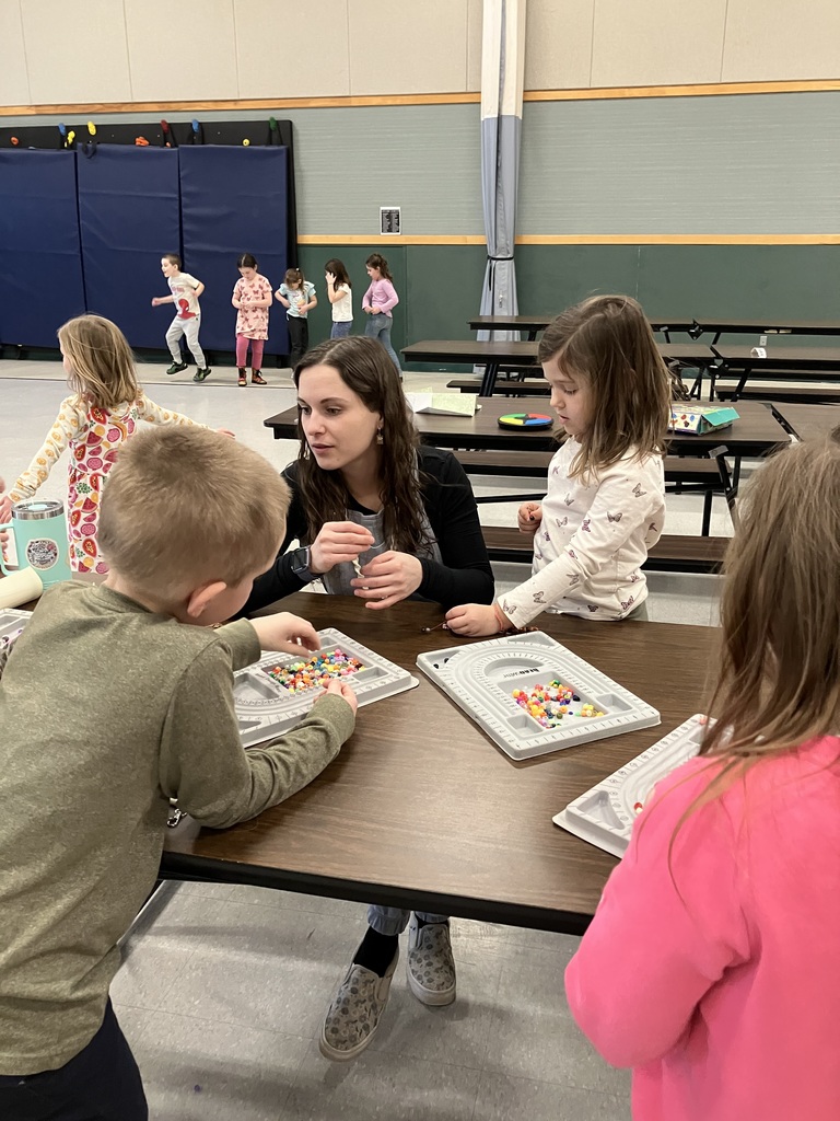 A woman and several young children sit at a long brown table, focused on stringing colorful beads using grey sorting trays. A young boy in a white heart-patterned t-shirt proudly holds up a finished string of beads while standing at a similar workstation.